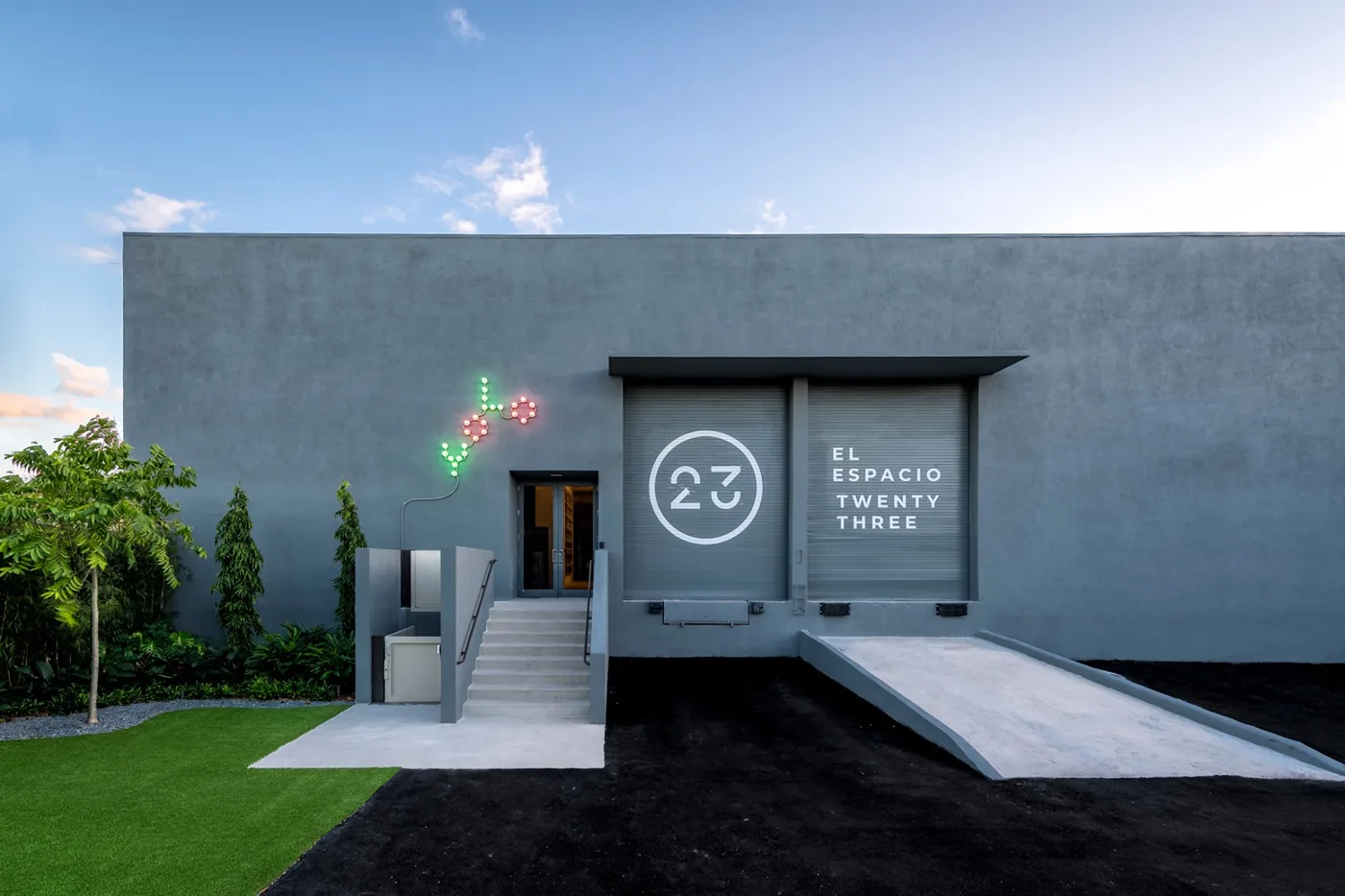 The gray warehouse facade of El Espacio 23 with its 23 logo on roll-up doors, a light sculpture on the wall and steps to the entrance