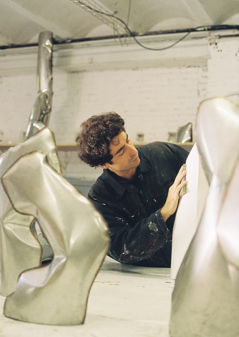 Gonzalo Guzmán working by hand on a curved stainless steel sculpture in a studio, with other steel forms around him