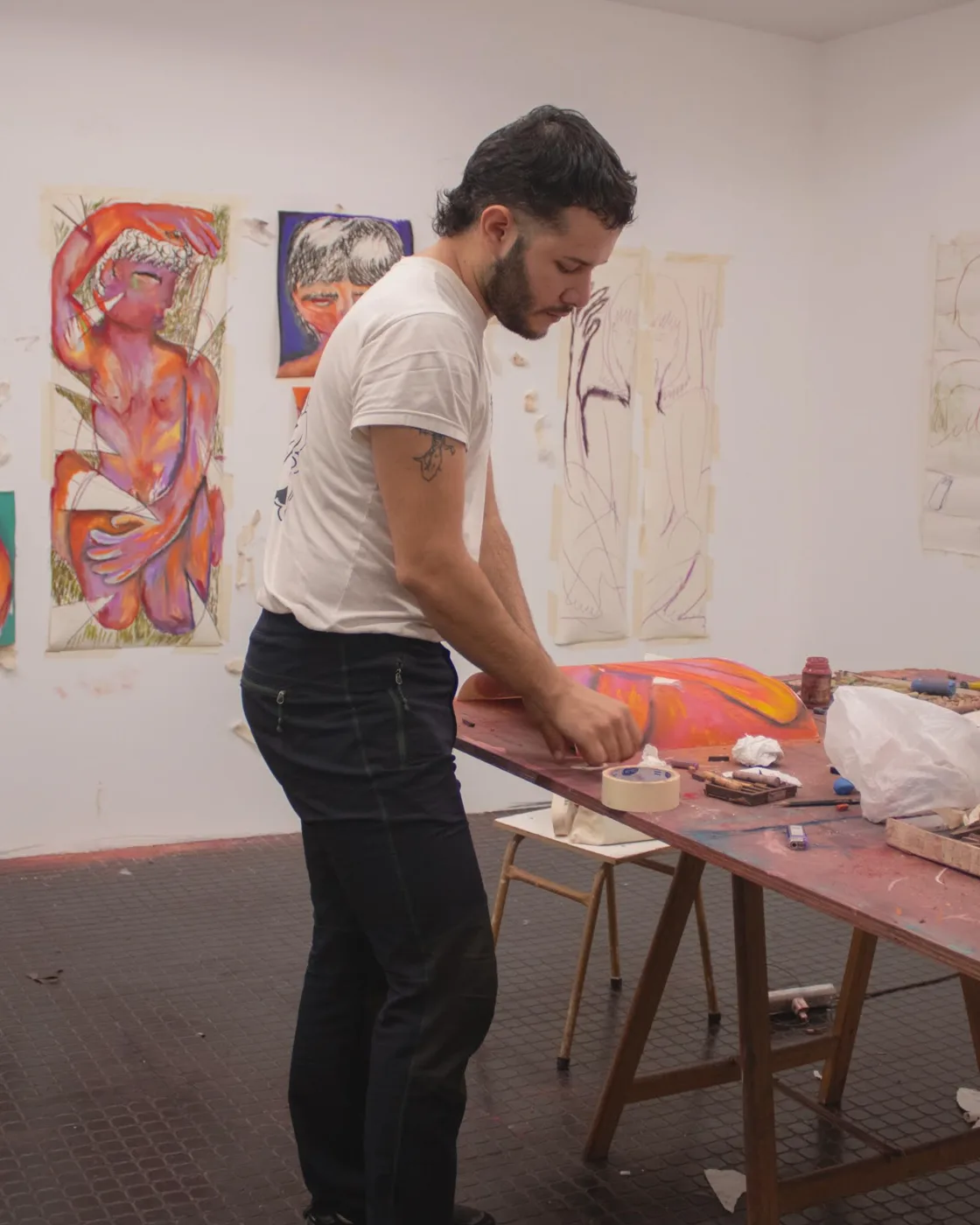 Ulises Mazzucca at a paint-covered worktable in a studio, with large colorful figure drawings taped to the walls