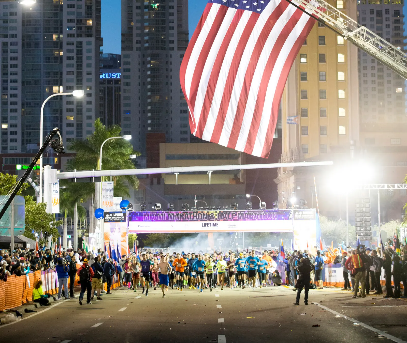 Runners leave the Miami Marathon start on Biscayne Boulevard under a flag hung from a fire ladder, with the Freedom Tower behind.